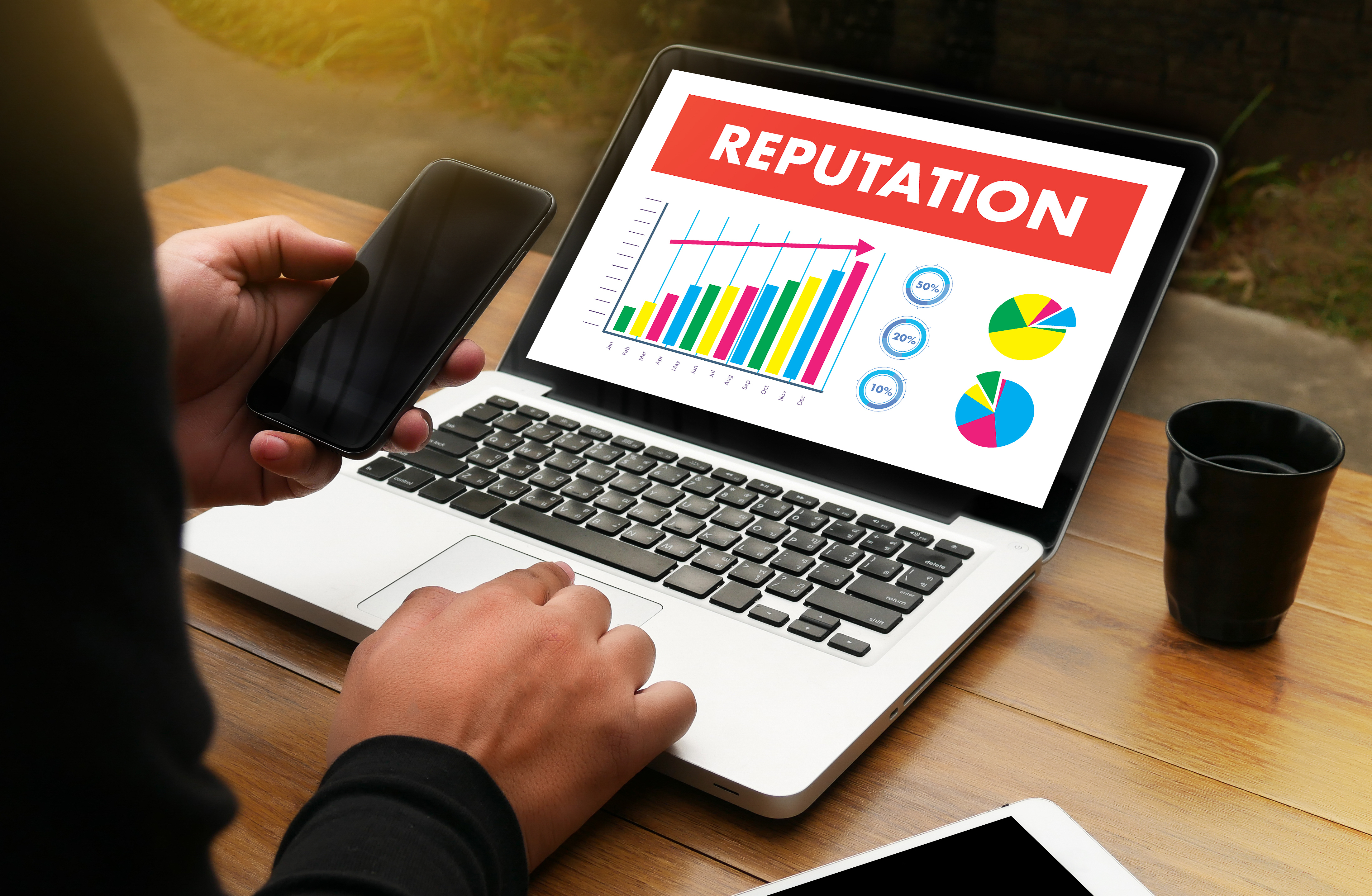 A picture of a person holding a cell phone and looking at a computer with the word reputation on the screen